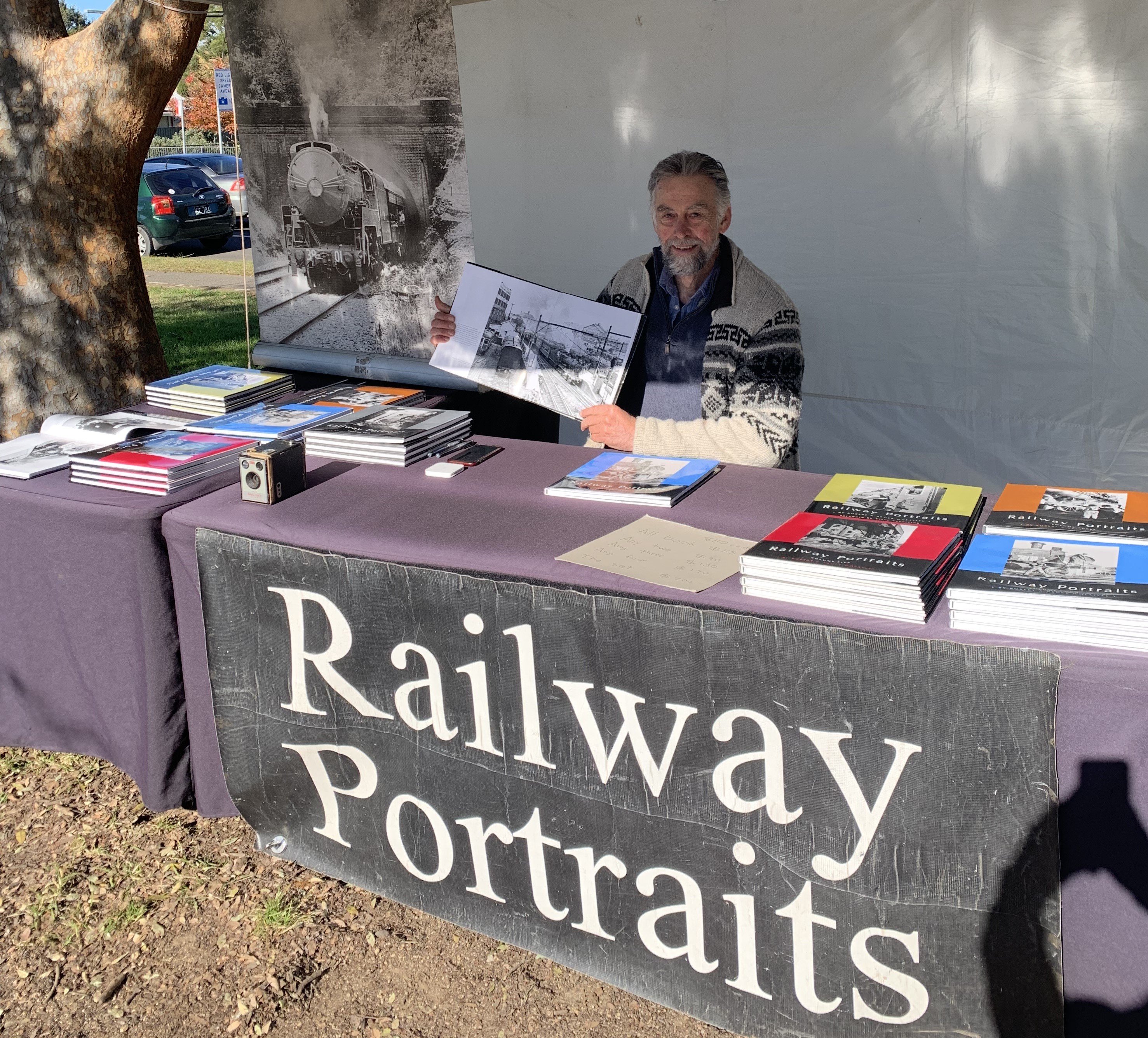 a photo of a man behind a market stall holding a photographic book. the front of the stall reads railways portraits, and atop the stall are several piles of coloured books with images of trains in black and white. behind the man sits a rectangular banner with a larger black and white image of a locomotive