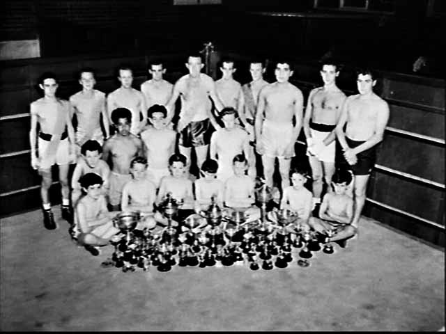 several men and boys sit on a boxing ring with trophies in front of them